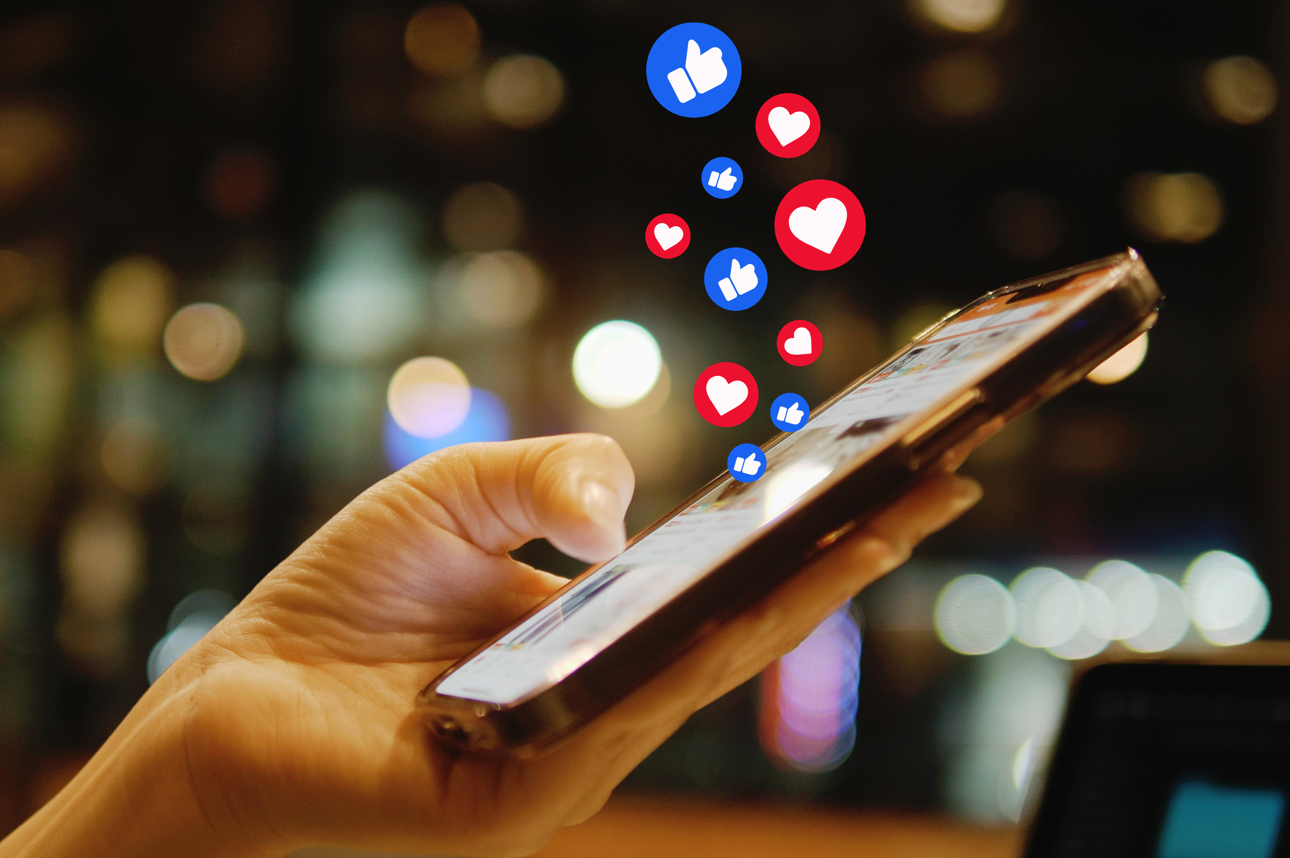 Hand holding a smartphone as red heart and blue thumbs-up reaction icons float up from the screen against a blurred city-lights background